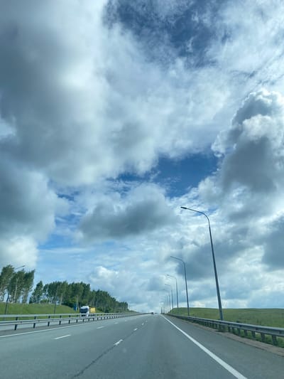 The image depicts a highway with a concrete road and guardrails. On the left side of the road, there is a line of trees and a white truck with a yellow trailer. The sky is filled with dramatic, dark clouds and patches of blue sky. On the right side of the road, there are rolling green hills and a series of lampposts. The scene appears to be a rural or semi-rural area, typical of the outskirts of a city like Vostochny, Russia. The time of day appears to be daytime, with the lighting suggesting it could be late afternoon.