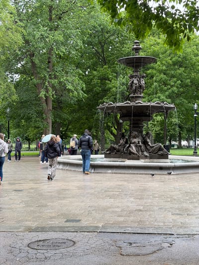The image depicts the Brewer Fountain, located in Boston Common, Boston, Massachusetts. The fountain is made of dark metal and features a multi-tiered design with sculptures and water flowing from each level. A group of people are standing near the fountain, seemingly observing or taking pictures. The scene is set on a paved area adjacent to the fountain, with trees and green foliage visible in the background. A sewer cover is also visible to the right of the fountain. The overall atmosphere appears to be a typical day in a public park.
