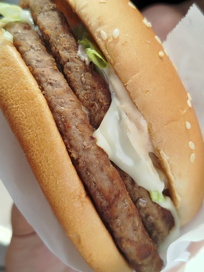 This image shows a close-up of a burger with two elongated meat patties in a sesame seed bun. Lettuce and a creamy white sauce are visible between the patties and the bun. The burger is held in a white paper wrapper. There is no information available in the image to ascertain that this image was taken in Noginsk, Russia.