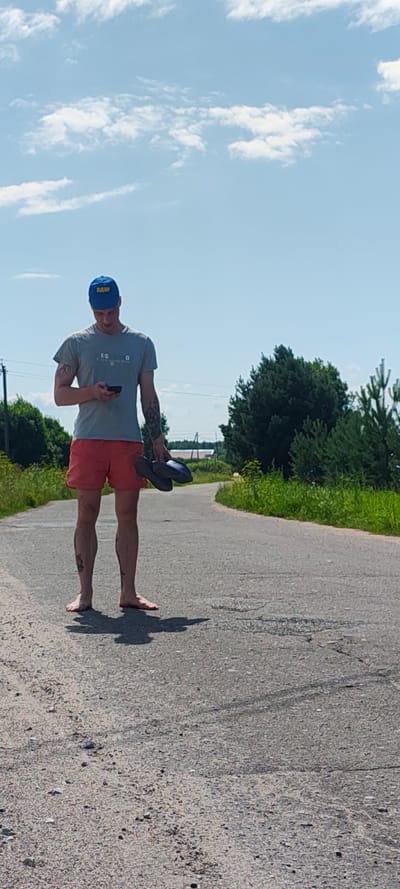 The image shows a young Caucasian male, wearing a grey t-shirt, red shorts, and a blue baseball cap with yellow lettering, standing on a paved road. He is holding a pair of dark-colored flip-flops in his left hand and looking down at his smartphone, which he holds in his right hand. The man is barefoot, and his shadow is visible on the road. The setting appears to be a rural area in Малая Листовка, Russia, with a paved road stretching into the distance, flanked by green trees and vegetation. The sky is blue with scattered white clouds. There are power lines visible in the background.