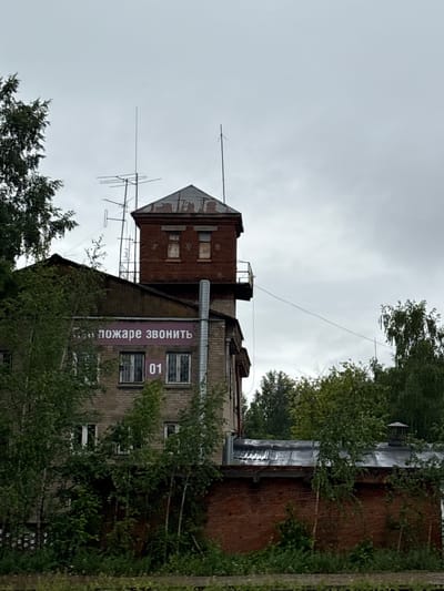 A medium shot captures a brick building in Krasnokamsk, Russia, under an overcast sky. The building appears to be old, with some signs of wear and tear. A prominent feature is a sign on the upper portion of the building that reads "ПОЖАРЕ ЗВОНИТЬ" (Call the Fire Department) in pink lettering. Below this, the number "01" is displayed in a larger font, likely indicating an emergency number. The building has several windows, some of which are boarded up. Vegetation, including trees and bushes, surrounds the structure, with some of it growing up against the walls, obscuring parts of the lower levels. Antennas are visible on the roof, suggesting a possible communication or monitoring function. The overall impression is one of an aging industrial or public facility.