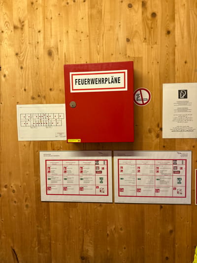This media file shows a wooden wall with various documents and a red fire safety box. The scene appears to be indoors, possibly in a public building or facility, given the presence of fire safety information. The wooden planks of the wall suggest a rustic or traditional aesthetic, and the lighting indicates daytime.

The most prominent object is a red fire safety box with the text "FEUERWEHRPLÄNE" (Fire Plans) prominently displayed in white lettering. Below and to the right of this box, a smaller, folded fire escape plan is attached to the wall. This plan is a diagram of a building layout with various symbols indicating exits, fire extinguishers, and other safety features.

To the right of the fire safety box, there are two large, multi-lingual safety posters. These posters, titled "NOTFALLPLAN" (Emergency Plan), appear to provide instructions in multiple languages, including German, English, French, Arabic, and Farsi, which aligns with the "Location context: Innsbruck, Austria" providing a diverse population. One poster also includes the logo "TIROLER SOZIALE DIENSTE" (Tyrolean Social Services). The text on these posters details emergency procedures and behavior in case of fire, with pictograms illustrating the instructions.

On the wall above the fire safety box, there is another document, partially visible, which seems to be a sign or information sheet, possibly with text in Arabic script. A small, circular "no smoking" sign is also visible near the top right of the fire safety box. The overall impression is one of an organized and safety-conscious environment. No people are visible in the image.