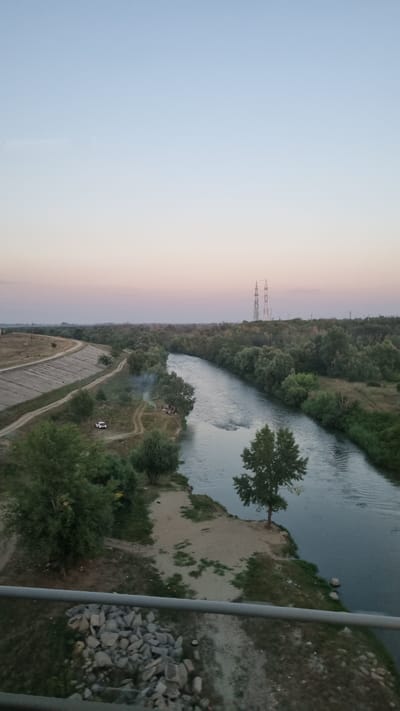 The image depicts a scenic view of the Argeș River as it flows through the commune of Adunații-Copăceni, Romania. The sky above is a soft gradient of pale blue, transitioning to a subtle pink and orange hue near the horizon, indicative of either sunrise or sunset.
On the left bank of the river, a paved embankment lines the water's edge, suggesting a man-made channel or flood control structure. A dirt path winds along this embankment, and a small white vehicle is parked near a group of people gathered around a smoking campfire. The surrounding vegetation consists of lush green trees and grasses.
The Argeș River itself is wide and appears to have a moderate current. The right bank is densely wooded with a variety of trees, creating a natural, untamed landscape. In the distance, two tall electricity pylons stand against the sky, a man-made intrusion into the otherwise natural scenery.
In the foreground, a gray metal railing runs horizontally across the lower portion of the image, suggesting the vantage point is from a bridge or elevated structure. Below the railing, a pile of large, irregular stones is visible, interspersed with dry grass and dirt. The overall impression is one of a tranquil, rural setting at the edge of a populated area, with the river serving as a central natural feature.