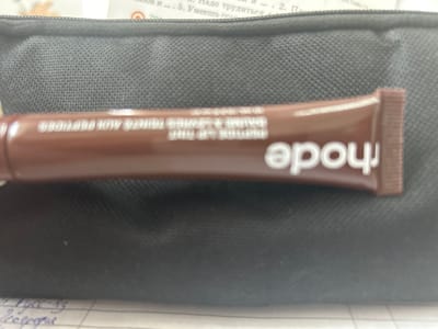 The image shows a brown tube of Rhode Peptide Lip Treatment lying on top of a black, textured pencil case. The tube is horizontally oriented with the word "rhode" visible in white lettering. "BAUME A LEVRES TEINTÉ AUX PEPTIDES" is printed above the brand name in smaller lettering. The top of a notebook with Cyrillic handwriting and text can be seen at the bottom left and top right of the frame. The image does not contain information about Dmitrievskaya, Russia, or any geographical location.