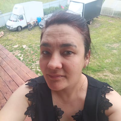 The image shows a woman taking a selfie outdoors, with a wooden deck in the foreground and a grassy yard behind her. In the background, there are several vehicles, including a white delivery truck, a motorcycle, and a black trailer. A greenhouse is also visible. The woman is wearing a black top with lace detailing on the shoulders. She has dark hair and is looking directly at the camera with a neutral expression. The scene appears to be set during the daytime, with overcast skies. The overall impression is of a casual outdoor setting, possibly a rural or suburban area. The location context is identified as Podlubno, Russia. There is no visible text in the image.