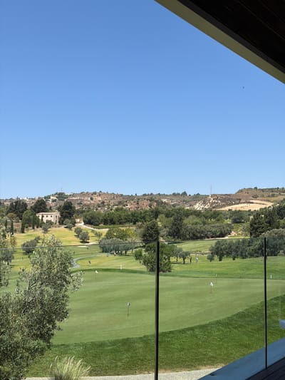 The image captures a sunny day on a golf course in Tsada, Cyprus, as viewed from a covered balcony with a partial wooden ceiling and a glass railing in the foreground. The expansive green fairways are dotted with white and black flags marking the holes. Several golfers are visible on the course, some walking and others possibly playing.

The landscape beyond the golf course features rolling hills covered in dry vegetation and scattered trees. A prominent stone building, possibly a historical structure or a clubhouse, is situated on one of the hills. In the distance, more buildings and a telecommunication tower can be seen on the crest of the hills, under a clear blue sky. The natural environment is characterized by Mediterranean flora, with olive trees and other greenery interspersed throughout the scene. The overall impression is one of a peaceful and well-maintained resort setting.