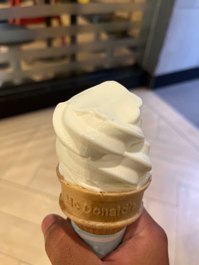 A close-up, hand-held perspective photograph shows a partially eaten McDonald's vanilla soft-serve ice cream cone. The soft-serve is swirled and conical. The waffle cone has "McDonald's" imprinted around the top. The cone is being held by a person's right hand, with only part of the hand visible. The background is blurred, with hints of neutral tones and metallic textures. The location and the city of Bencongan Indah, Indonesia cannot be determined from the image.