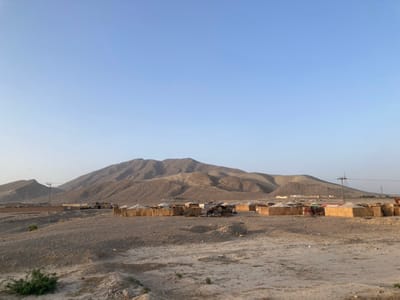 The image displays an arid landscape in Spezand, Pakistan, under a clear, light blue sky. A range of treeless, light brown mountains with visible geological contours dominates the background. In the midground, an informal settlement extends across the dusty, light brown terrain. This settlement comprises numerous single-story structures, many constructed from light-colored, woven materials such as reeds or straw, possibly with canvas or tarp elements. Some structures also exhibit more solid, possibly mud-brick, construction. These dwellings are clustered without a defined grid, interspersed with scattered debris and uneven ground. Several utility poles with overhead power lines traverse the scene horizontally across the midground. The foreground consists of rough, sandy, and rocky ground, with a small patch of sparse green vegetation visible in the bottom left corner. No individuals or discernible activities are present. The scene is illuminated by natural daylight.