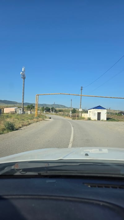 The image depicts a rural roadside scene under a clear blue sky, presumably in or near Stalskoye, Russia, given the prompt's context. A paved road curves to the right, with a white dashed line marking the center. On the left side of the road, a tall cell tower stands prominently. A yellow pipeline arches over the road, supported by poles. To the right of the road, there is a small, white, cubical building with a blue roof and an open doorway. In the foreground, the dashboard and windshield of a vehicle are visible, indicating the perspective is from inside a car. The surrounding landscape consists of dry grass, scattered bushes, and distant hills or mountains. Several utility poles and fences are also present in the scene.