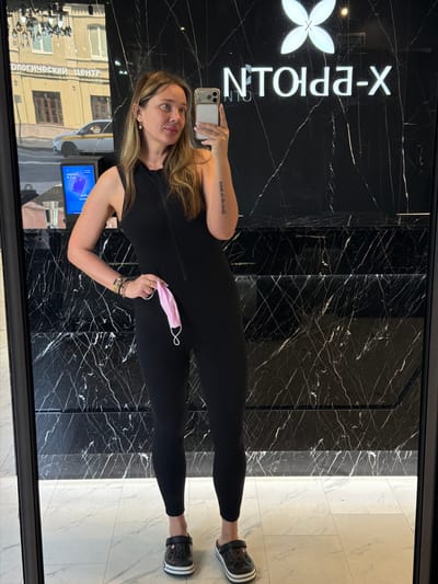A woman stands in front of a mirrored wall, taking a selfie. She is wearing a black sleeveless jumpsuit and black Crocs. She is holding a pink face mask in her right hand. In the reflection of the mirror, we see a street scene with buildings and a white car. The wall behind her is black marble with white veining. A logo reading "NTOLPA-X" is visible on the wall. The floor is tiled in a light color. The image appears to be taken indoors, likely in a salon or shop.