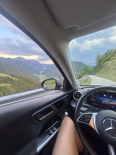 The image is a first-person perspective from inside a car, driving on a scenic mountain road. The car's interior is visible, including the steering wheel, dashboard displaying information like speed and range, and the car door with window controls. Outside the window, a dramatic landscape of steep, forested mountains unfolds, with a valley containing a village visible in the distance. The sky is partly cloudy with a warm, late afternoon light. The road itself is paved and winds upwards, bordered by a guardrail and steep, grassy slopes with trees. The location is specified as Ratschings - Racines, Italy, suggesting the scene is set in the South Tyrolean Alps. A person's leg is visible in the foreground, indicating they are the passenger or driver. A bottle of water is also present on the car door.