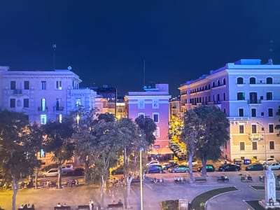 The image captures an urban scene at night, likely from an elevated perspective. The sky is a deep, dark blue, indicating nighttime. In the foreground, a paved area with curved green borders is visible, with several people seated on benches. A tall, slender monument or sculpture stands to the right, with a single person seated at its base. Beyond this, trees with dark foliage line the area, with some illuminated by streetlights.

In the mid-ground, a street is lined with vehicles, including a white sedan, a blue hatchback, and several dark-colored cars, one of which is a dark green SUV. Buildings with illuminated windows and architectural details are prominent. On the left, a light pink building features multiple floors with windows and balconies. To its right, a bright pink building has a prominent window on its upper level and lights on its lower floors, suggesting commercial establishments. Further to the right, a cream-colored building exhibits a row of arched windows on its upper floors and balconies. Several streetlights illuminate the area.

The background consists of more buildings, some with illuminated windows, and the dark night sky. The overall impression is of a city square or street scene at night, with a mix of residential and commercial buildings, public spaces, and vehicular traffic. The image does not contain any elements that suggest the location is Porrentruy, Switzerland.