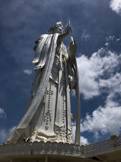 This media file shows a low-angle, upward view of a large, white, ornate statue against a bright blue sky dotted with white clouds. The statue appears to be made of marble or a similar material and depicts a figure with flowing robes and elaborate detailing. One of the statue's hands is visible, extended forward, holding a cylindrical object. Another hand is visible reaching out. The statue is set against the backdrop of a building with decorative architectural elements, suggesting it might be part of a temple or religious site. The overall impression is one of grandeur and artistry, with the bright sunlight highlighting the intricate details of the sculpture. The scene is likely set during daytime, given the strong sunlight and clear sky. The location is identified as Nam Ban, Vietnam. There is no visible text in the image.