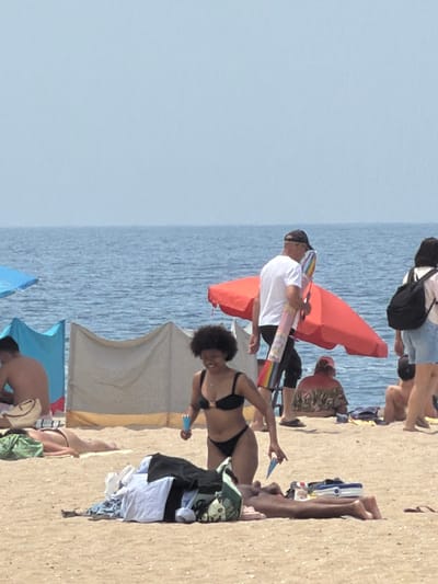 The image depicts a sunny day at a beach, likely in Espinho, Portugal, given the context of a previous request. In the foreground, a woman with dark, curly hair is wearing a black bikini. She is holding a blue object, possibly an ice pop or a toy, in her right hand and reaching towards the sand with her left. A pile of clothes lies on the sand in front of her. Behind her, another person is lying on the sand with a green towel draped over their legs. Further back, a man in a white t-shirt and black shorts, wearing a dark baseball cap, is walking and carrying a rolled-up colorful item under his arm. He is near a large red umbrella. To the left, a blue beach umbrella is partially visible. In the background, the blue ocean stretches to the horizon under a pale blue sky, with several other beachgoers and beach shelters scattered across the sandy shore.