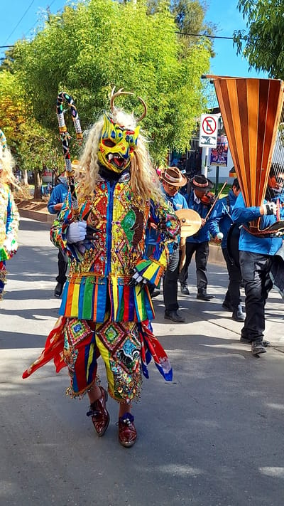 A person in a vibrantly colored, elaborate costume and mask is walking in a street parade in Wanchaq, Peru. The mask is yellow with red and blue accents and a long, blonde wig. The costume is adorned with numerous colorful beads, sequins, and fabric strips. The person is holding a decorated staff in their left hand. Behind them, other individuals are visible, some playing musical instruments, including a harp-like instrument and a violin. They are dressed in blue shirts and dark pants, and some are wearing hats. The ground is asphalt, and trees and buildings are visible in the background. The sky is clear and blue. A circular sign with a red border and a black 'E' symbol is visible on the right side of the frame.
