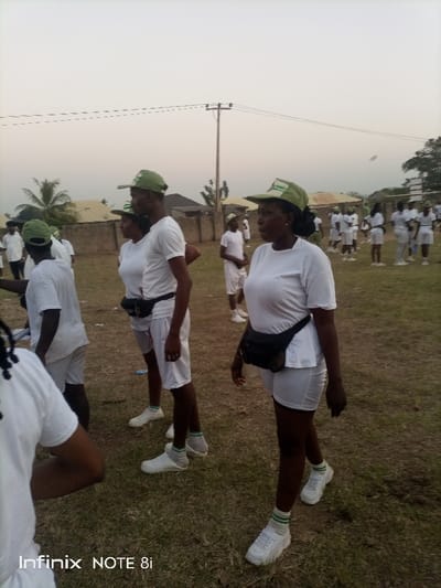 A group of people are wearing white t-shirts and shorts with green caps in an outdoor setting. They are standing on a grass field with a utility pole visible in the background. The people are wearing black fanny packs and white sneakers with green and white striped socks. Some of the people are standing in a group while others appear to be walking. The overall setting suggests a gathering or event, possibly a training exercise or organized activity.
