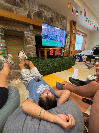 In Washington Township, United States of America, a group of young adults are gathered in a room, likely a common area or recreational space, to watch a soccer match on television. The room features a prominent stone fireplace with a wooden mantel. A large flat-screen television is mounted above a wooden table, which is covered with artificial green turf. The television displays a soccer game with a stadium filled with spectators.
Several individuals are seen lounging comfortably, with one person lying on their back on a large gray beanbag chair, looking up at the TV. This person has a bandage or face paint, possibly representing the English flag, on their forehead. Another person is lying on the floor with their legs crossed, wearing shorts and a blue t-shirt, with a phone placed on their stomach. Two other individuals' legs are visible in the foreground, one wearing athletic shoes and socks, and another wearing Crocs. The floor is a mosaic of yellow and orange tiles.
The room is decorated with a string of small flags, likely representing various countries participating in a tournament. A wooden cabinet with multiple plaques is mounted on the wall to the right of the television. In the background, other furniture and objects, including chairs and what appear to be large cardboard cutouts, are visible. String lights are draped near the floor, adding a festive atmosphere. The overall scene depicts a relaxed and engaged gathering of friends enjoying a sporting event.