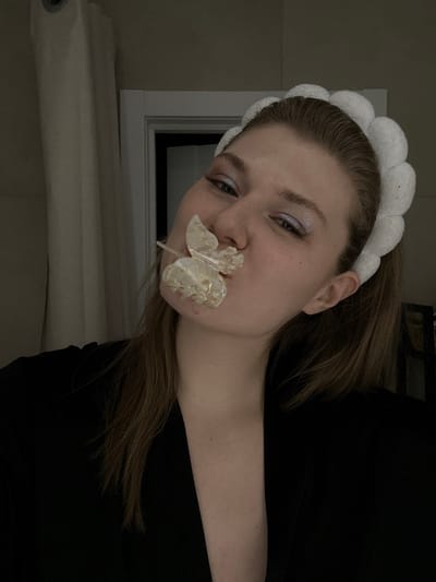 A young woman tilts her head toward the camera. She wears a white, puffy headband and has fair skin with light makeup. A large, translucent butterfly with a stick attachment is held in her mouth. She wears a black top. The background includes a white window frame and a white rolled-up object, possibly fabric, leaning against the wall. The lighting is dim.