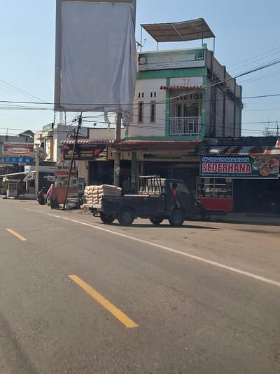 This image depicts a street scene in Meunasahpulo, Indonesia, with buildings and a road in the foreground. On the left side of the road, a store with a blue and red "Indomaret" sign is visible. Further down the street, there is a "Toko Mas" (gold shop) building with a white banner draped over its upper section. Adjacent to it, a building displays a "Rumah Makan Sederhana" (simple restaurant) sign. In the middle of the scene, a black pickup truck is parked, carrying stacks of bagged goods, likely cement, on its bed. A person is visible inside the truck's cabin, and another person in a pink headscarf is walking near a food stall on the sidewalk. The road itself is paved with asphalt and marked with yellow and white lines. The sky is clear and blue.
