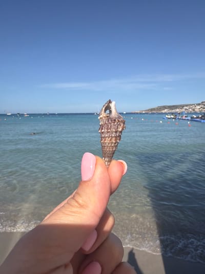A close-up shot focuses on a person's hand holding a small, ribbed, brown and white seashell. The hand has light skin and neatly manicured pink fingernails. In the background, the turquoise waters of a beach stretch towards the horizon under a clear blue sky. Small boats and a floating platform are visible on the water. The shoreline is lined with orange buoys. The scene appears to be at Għadira Bay in Mellieħa, Malta.