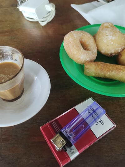 An overhead shot shows a table setting with a glass of coffee, a plate of fried pastries, and a pack of cigarettes with a lighter. The glass of coffee is half-full and sits on a white saucer. On a green plate, there are two ring-shaped donuts and a fried spring roll. A pack of red and white cigarettes lies in the foreground, with a clear plastic lighter resting on top. The objects are on a wooden table. The location is Keudematangkuli, Indonesia.