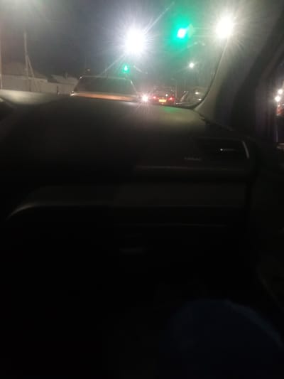 The image depicts a view from inside a car at night, likely in traffic, with a green traffic light visible. The dashboard of the car is in the foreground, with the word "AIRBAG" visible on it. Beyond the car's windshield, other vehicles are present, their headlights and taillights creating streaks of light. The overall scene suggests a nighttime urban environment, possibly a city street with traffic lights. The bright lights in the image, particularly the green traffic light and the headlights of other cars, indicate that it is dark outside, consistent with nighttime. There are no clear indications of weather conditions, but the bright lights suggest a clear or mildly hazy night. The setting appears to be a road or intersection where traffic is present. No people are clearly discernible as subjects.