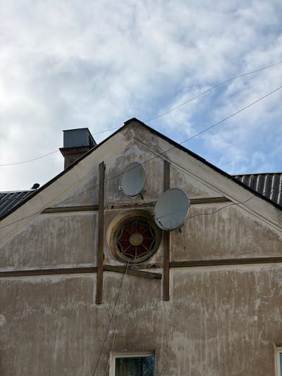 A low-angle shot depicts the exterior of a building, possibly in the city of Tokkari, Russia, based on the instructions. The building's architecture includes a pointed gable with two satellite dishes mounted on it. There is also a round, ornate window with a reddish-brown design. The building's facade is textured and aged, with visible signs of wear and tear. Wooden supports are visible, adding to the building's structure. The sky is visible in the background, with some clouds. A power line is visible crossing in front of the building. There are some black tiles on the roof.