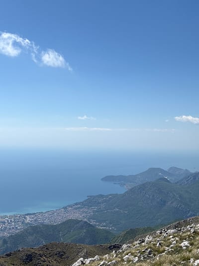 The image shows a view from a high vantage point in Mali Mikulići, Montenegro. The scene includes a body of water, likely a lake or part of the Adriatic Sea, a town or city built along the coast, and the rugged terrain of the mountains. The foreground includes rocky, grass-covered terrain. The sky is a clear blue with a few scattered clouds. The overall impression is that of a scenic landscape viewed from a high altitude.
