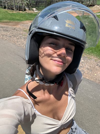 A woman wearing a grey helmet with a gold "Royal" emblem and a clear visor is looking directly at the camera. She is wearing a light-colored, sheer long-sleeved top over a ribbed white tank top and denim shorts. Around her neck, she has a necklace made of white shells. She is outdoors on a paved road, with a grassy area and some wooden structures in the background. The scene appears to be in a rural or semi-rural area.