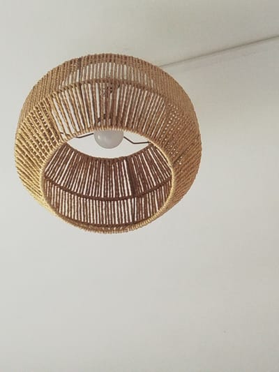 A close-up, low-angle shot shows a pendant light fixture made of natural-colored rope, suspended against a white ceiling. The light fixture is spherical in shape with an open bottom, revealing a bare lightbulb inside. The rope is woven vertically and horizontally, creating a textured, basket-like appearance. The background is predominantly a plain white ceiling with a subtle shadow or seam visible on the upper right side. There are no people or other objects in the frame. The image appears to have been taken indoors, likely in a residential or commercial space. The location in Saint-Claude, France, is not discernible from the visual information provided.
