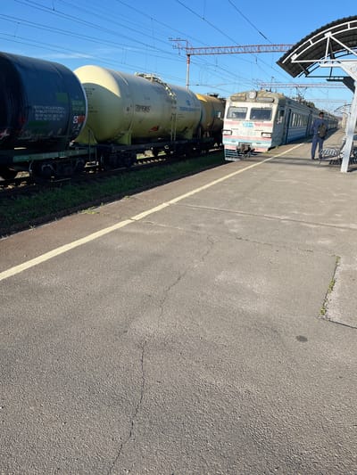 This is a daylight, eye-level, medium shot of a train at a station in Kalynivka, Ukraine. In the foreground, a paved train platform is visible with a white line marking its edge. To the left of the platform are two large, cylindrical tank cars on a train track. The first tank car is dark gray and the second is a pale yellow. Both cars have visible markings and text. A modern electric train, primarily white and light blue with red accents, is stopped at the platform. The train has the designation "ЕР2-384" and a "УЗ" logo on its front. A person wearing dark blue trousers and a dark top is standing on the platform facing the train and looking away from the camera, holding a black backpack. In the background, overhead power lines and a station roof structure are visible. The sky is clear and blue.