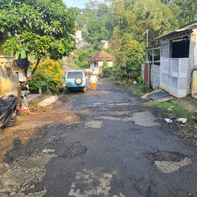 The image depicts a narrow, paved street in Kecamatan Tamansari, Indonesia, at coordinates -6.635 latitude and 106.738 longitude. The street is in poor condition, with numerous potholes and uneven surfaces. A blue minivan is parked on the right side of the street. A man wearing a long orange robe is walking toward the camera, away from the minivan. On the left side of the street, there is a parked motorcycle and foliage. There are also some birds on the street in the distance. In the background, there is a house on the right side of the street and a small building on the left side of the street. The overall environment is rural, with lush greenery surrounding the street.