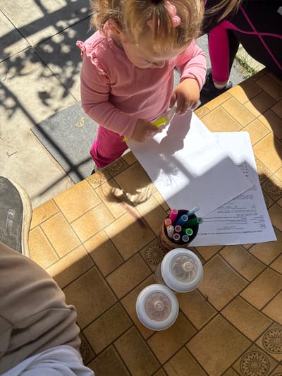 Here is a detailed description of the image:
**Scene:** An overhead shot showing a section of a patterned table where a young child is seated, coloring on a piece of white paper.
**People:** A young child with blonde hair, wearing a pink shirt and pink pants, is holding a yellow marker. There is another person nearby wearing a black and pink outfit. A third person is wearing beige pants and a white shirt.
**Objects:** There are documents on the table. Near the child is a piece of white paper which she appears to be coloring on. Beside the paper is a container of colorful markers. Two containers with lids are on the table.
**Actions/Interactions:** The child is engaged in coloring or drawing on the paper. The other people are sitting around the table.
**Environment:** The image appears to be taken either outdoors or in a sunlit area, as indicated by the shadows cast on the table and the presence of gray concrete or stone tiles underneath a portion of the table.
The OCR indicates the city is Eline Pelin. Baylovo is not referenced.