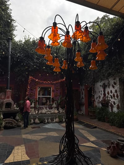 A vibrant outdoor scene features a black metal sculpture with orange, flower-shaped lights. Behind it, a building decorated with intricate designs and vines adds to the artistic ambiance. A row of white pots with plants lines the walkway. A person in a pink shirt and jeans is seen near the pots. The pavement displays a colorful, geometric pattern. The sky appears overcast.