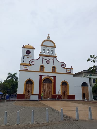 The image shows the exterior of a white church with yellow and red accents in Tolú, Colombia. The church has a central bell tower with two bells, flanked by smaller towers. The facade features arched wooden doors and windows, some adorned with decorative ironwork. A cross is visible on the central tower and above one of the arched windows. In front of the church, there is a paved plaza with some bollards. A ladder, paint cans, and a person in a yellow vest are present near one of the large wooden doors, suggesting ongoing maintenance or painting work. Several palm trees and other vegetation are visible to the left and right of the church, along with a glimpse of other buildings in the background. The sky is overcast.