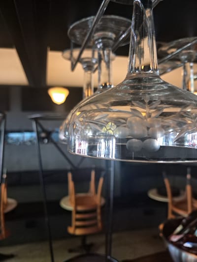The image depicts a close-up of a row of wine glasses with etched floral designs, possibly arranged on a drying rack, in what appears to be a commercial setting. The background shows blurred chairs and lighting, suggesting a restaurant or bar environment. The location is specified as Vicente López, Argentina.