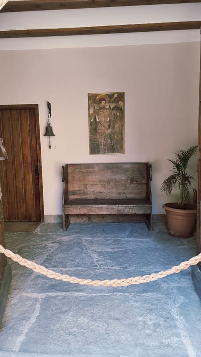This is a full, eye-level, vertical shot of an entryway or courtyard in Rupit i Pruit, Spain. The scene features a rustic wooden bench with a weathered finish positioned centrally against a pale pink wall. Above the bench hangs a framed painting depicting two figures in historical attire. To the left of the bench is a tall, wooden door with vertical planks. A small, dark-colored bell hangs by a chain from a bracket on the wall to the left of the door. To the right of the bench, a green plant in a terracotta pot sits on the stone floor. The floor is made of large, irregularly shaped stone slabs, giving the impression of an old or natural setting. A thick, braided rope barrier runs horizontally across the lower portion of the image, separating the viewer from the depicted scene. Wooden beams are visible along the top of the frame, suggesting an overhead structure like a porch or covered walkway. The lighting is bright and natural, indicating daytime.
