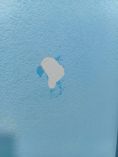 The image is a close-up, eye-level shot of a textured, light blue wall in what appears to be an indoor setting. The wall has a rough, uneven surface with visible bumps and indentations. There is a patch of chipped or peeling paint in the center of the frame. The exposed area beneath the chipped paint is a lighter, off-white color, and some areas show a brighter blue underneath. There are also some small, lighter-colored splotches on the wall in the upper left quadrant, possibly indicating discoloration or a different type of paint. The lighting is somewhat diffused, creating soft shadows and highlighting the texture of the wall. The image does not provide any specific geographical markers or context to indicate its location in Tokmok City, Kyrgyzstan. There are no people, animals, or other objects visible in the scene.