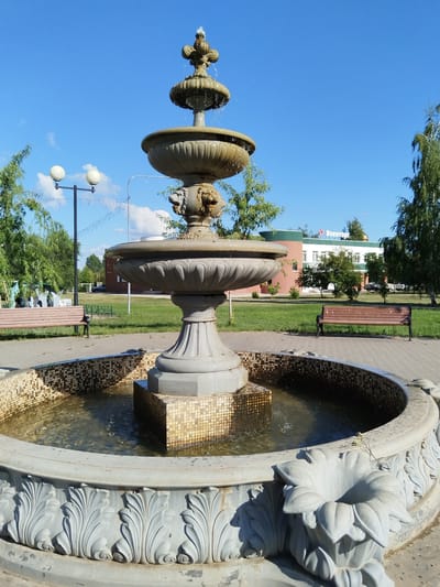 This is a photograph of a multi-tiered fountain in a park setting. The fountain is made of stone and features ornate carvings, including what appear to be lion heads on the lower basin. Water is flowing from the top tier, cascading down into the lower basins. The fountain is surrounded by a circular pool with a mosaic tile border. In the background, there is a paved walkway, a grassy area, trees, benches, and a building with Russian text and a red star logo that reads "Военторг" (Voentorg). A lamppost with two round lights is also visible. The sky is blue with some scattered white clouds. The image appears to be taken on a sunny day. This scene is located in the city of Roshchinsky, Russia.