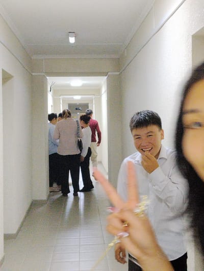 The image depicts a hallway scene with several people. In the foreground, a young man with short dark hair is smiling and holding his hand near his mouth, appearing to be mid-bite. He is wearing a white collared shirt. To his right and partially out of frame, a person's hand is held up, forming a peace sign, adorned with rings and painted fingernails. In the background, a group of people are gathered at the end of the hallway, some facing away from the camera. They are dressed casually, with one person in a light blue shirt, another in a light pink sweater and dark pants, and another in a dark red shirt. The hallway has white walls and a tiled floor with a light-colored pattern. Overhead, fluorescent lights illuminate the space. The scene appears to be indoors, possibly a school or residential building hallway. The lighting suggests it could be daytime, though no windows are visible. The overall mood seems casual and friendly, with the smiling subject in the foreground and the peace sign gesture. No text is visible in the image.