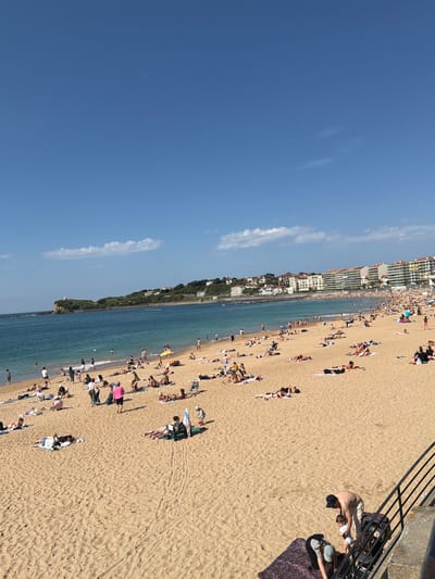 This photograph shows a beach in Saint-Jean-de-Luz, France. The beach is crowded with people sunbathing, swimming, and engaging in various beach activities. Buildings line the coast behind the beach. A metal railing is visible in the foreground on the right side of the image. A man wearing light-colored pants, a dark hat and no shirt is shown kneeling to attend to another person.