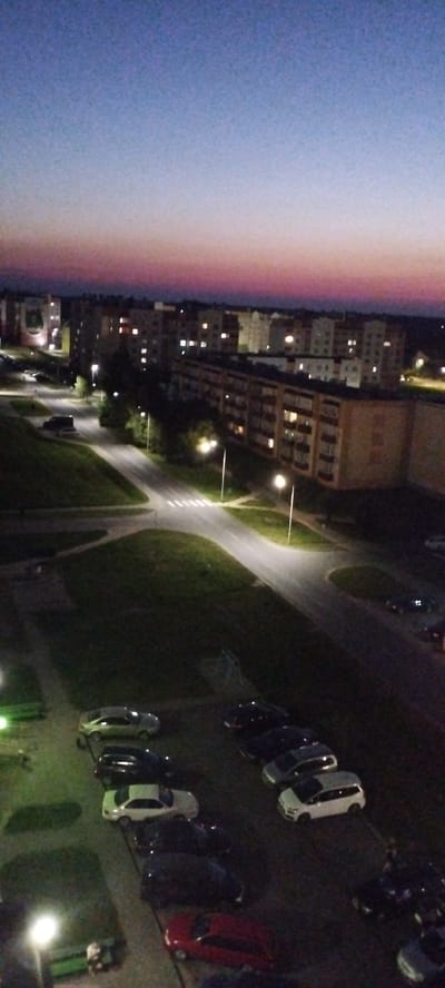 This is a vertical, medium-high angle, nighttime photograph of a residential area at dusk in Skidzieĺ, Belarus. The sky exhibits a gradient of colors from deep blue at the top to pink and purple near the horizon. In the background, numerous apartment buildings are visible, with many windows illuminated. A multi-lane road with streetlights dissects the foreground and midground, curving through a grassy area and intersecting with other paths. Parked cars of various colors are scattered throughout the scene, particularly in parking lots and along the roadside. The lighting suggests it is late evening or early morning, with artificial lights from buildings and streetlamps providing illumination.