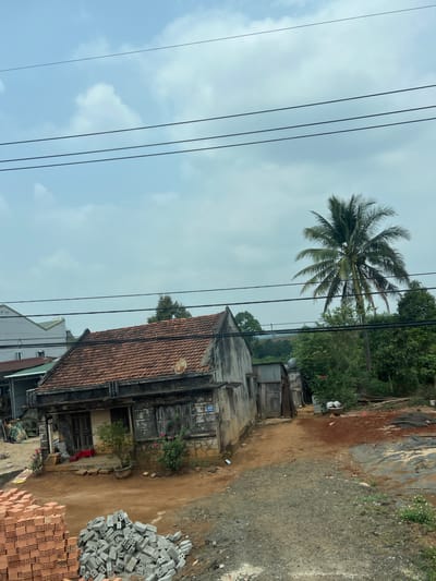 The photograph shows an exterior view in Thong Nhat Ward, Vietnam. A weathered, traditional building with a tiled roof is centrally positioned. To the side are piles of construction bricks and stones, suggesting ongoing or planned construction. Overhead power lines are visible against a cloudy sky. A tall palm tree stands in the background amidst other vegetation. The surrounding area appears to be a mix of older structures and natural landscape. The ground is dirt and gravel.