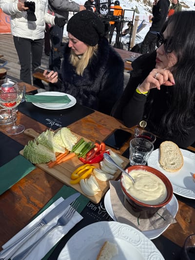 Two women are seated at a wooden outdoor table, enjoying a meal or drinks on a sunny day in a snowy mountain setting. The image is oriented vertically with the table angled diagonally across the frame.
The woman in the foreground on the left has blonde hair, wears a black beanie and a dark faux fur jacket, and is looking down at a smartphone in her hands. To her right, a woman with long dark hair and sunglasses is visible from the chest up. She wears a black top, has a silver ring on her left ring finger, and appears to be looking towards the table. Other individuals in winter clothing are partially visible in the snowy background, suggesting a ski resort environment.
On the table, a spread of food is laid out. A large brown bowl holds a creamy white dip, possibly fondue or a similar sauce, with a spoon resting in it. Beside it, slices of white bread are placed on a decorative white plate. A wooden serving board features a colorful array of raw, sliced vegetables including red and yellow bell peppers, celery sticks, carrot batons, sliced fennel, and two piles of chopped savoy cabbage or lettuce.
Two wine glasses are visible, one containing a red liquid and another clear glass. White plates, silverware (a fork and knife), and green napkins are arranged on the table. A black smartphone is also resting on the table near the dark-haired woman.
Visible text on a black placemat or menu item reads "ALPEN TOP" with a logo resembling a deer or ibex head, followed by "ALAGNA" and partially "MONTE RO**", confirming the location as Alagna Valsesia, Italy, as provided in the context. The bright daylight casts distinct shadows, indicating a clear, sunny sky, likely midday or early afternoon. The overall atmosphere appears relaxed and social in a winter mountain resort.