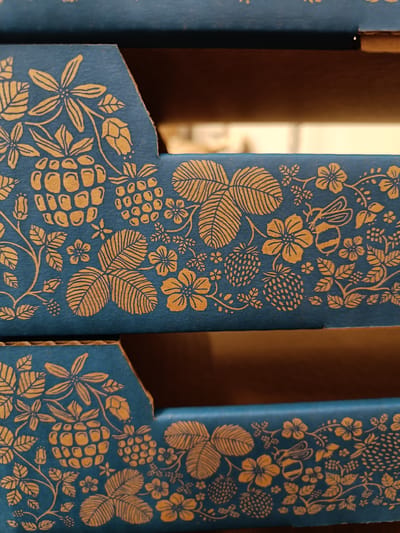 The image shows a close-up of two stacked blue cardboard boxes with a decorative pattern of golden-yellow strawberries, leaves, flowers, and a bee. The pattern is intricate and appears to be printed or embossed onto the surface of the boxes. The boxes are positioned at an angle, suggesting they are part of a larger display or packaging. The background is blurred, but appears to be the interior of another box or a dark space. The lighting is even, suggesting indoor lighting. No people are visible. The overall impression is of a decorative and possibly handcrafted item. The location context is Tuusniemi, Finland.