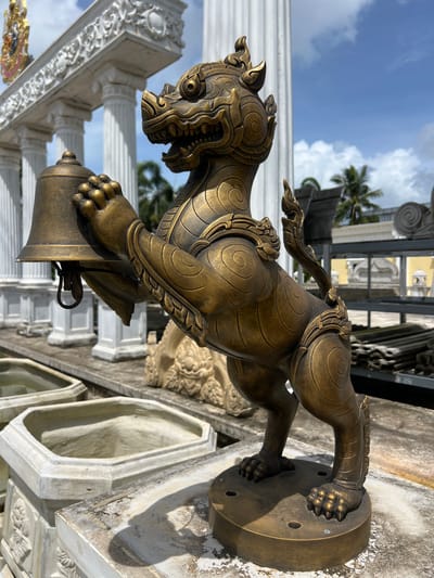 This media file features a striking bronze statue of a mythical creature, possibly a Foo dog or a Kirin, with intricate detailing and a golden-brown patina. The statue is positioned outdoors, with a bright blue sky dotted with white clouds visible in the background, suggesting a sunny day. Palm trees and hints of elaborate architecture, likely a temple or ornate building with white columns and decorative carvings, are also present, indicating a tropical or Southeast Asian setting. The statue itself is depicted in a dynamic pose, with its front paws resting on what appears to be a bell or a similar decorative object. The creature has a fierce expression with sharp teeth and stylized facial features. The statue is mounted on a circular base, which is screwed into a weathered concrete surface. The overall scene conveys a sense of grandeur and cultural significance, common in religious or historical sites. The image is captured from a low angle, emphasizing the scale and presence of the statue. No people are visible in the frame, and there is no visible text. The lighting suggests it is daytime, likely late morning or early afternoon, given the strong shadows cast by the statue. The setting in Pa Khlok, Thailand, further supports the interpretation of a culturally rich and potentially religious location.