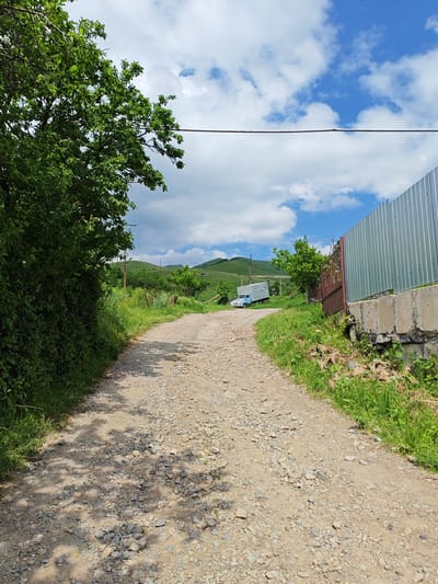 The image depicts a dirt road winding through a rural landscape. On the left side of the road, dense green foliage and trees cast shadows on the path. To the right, a corrugated metal fence runs along a concrete and stone wall, bordering the property. Beyond the fence, a rusty metal gate is visible, along with a small blue truck parked on the side of the road. The road continues uphill, leading towards rolling green hills under a partly cloudy blue sky. The scene appears to be located in the city of Bazum, Armenia, judging by the context provided.