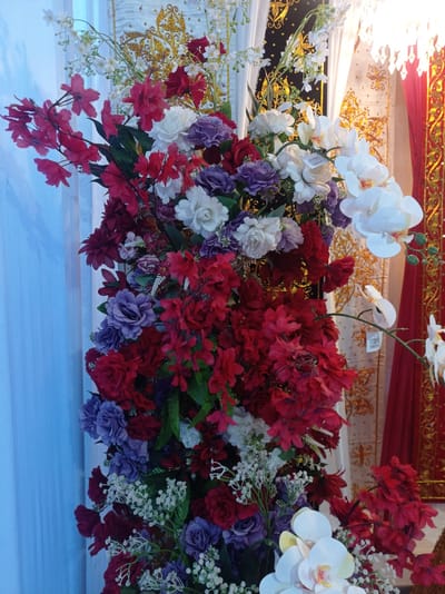The image displays an arrangement of artificial flowers, predominantly in shades of red, purple, and white, with some smaller white flowers and greenery. The floral arrangement is lush and dense, filling much of the frame. In the background, draped fabric can be seen, with a white fabric adorned with gold patterns and a section of red fabric with gold trim. A crystal-like chandelier is partially visible in the upper right corner, suggesting an indoor setting, possibly a venue for an event. The presence of the gold-patterned fabric and the overall opulent aesthetic hint at a decorative display, perhaps for a wedding or other celebration. Without further context, it is not possible to ascertain the specific location in Peudari, Indonesia, beyond it being an indoor decorated space. There are no people or animals present in the image.