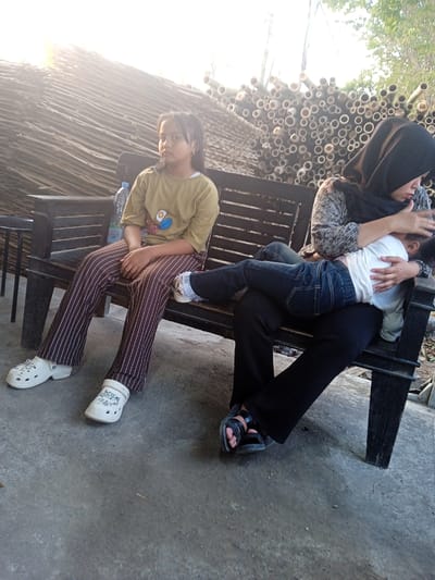 Two young girls and a woman are seated on a dark wooden bench. A young girl with long dark hair and a mustard-colored shirt with a graphic is sitting on the left side of the bench, facing forward. Her legs are crossed at the knees, and she is wearing brown and white striped pants and white clogs. To the right of her, a woman wearing a black hijab and a patterned top is holding a younger child who is lying across her lap, facing away from the camera. The child is wearing a white t-shirt and dark blue jeans. The woman is wearing black pants and black sandals. The bench is on a concrete surface. In the background, there is a stack of cut bamboo poles and a roof structure made of wooden planks. The scene appears to be outdoors, possibly in a rural or semi-rural area of Punden, Indonesia, given the presence of bamboo and the natural lighting.