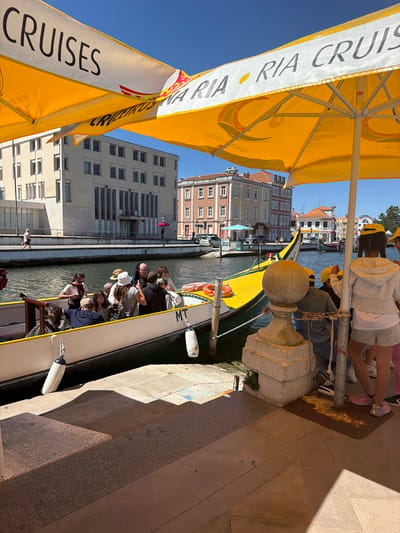 The image depicts a sunny day scene along a canal in Aveiro, Portugal, likely near the Praça da República or a similar central waterfront area. Yellow umbrellas with the text "CRUISES" and "RIA CRUISE" are prominent in the foreground, casting shadows. A traditional Moliceiro boat, painted in bright yellow with a decorative prow, is docked and filled with tourists. The boat has the inscription "MT" on its side. Passengers, including adults and children, are visible in the boat, some looking out at the canal and others interacting with each other. On the canal banks, a pedestrian walkway with buildings lining it can be seen. The architecture suggests a historic Portuguese city, with buildings featuring colorful facades and shuttered windows. People are walking along the promenade, and cars are visible in the background. The water in the canal is calm, reflecting the blue sky. The overall atmosphere is one of a tourist attraction, with leisure activities and a lively ambiance.